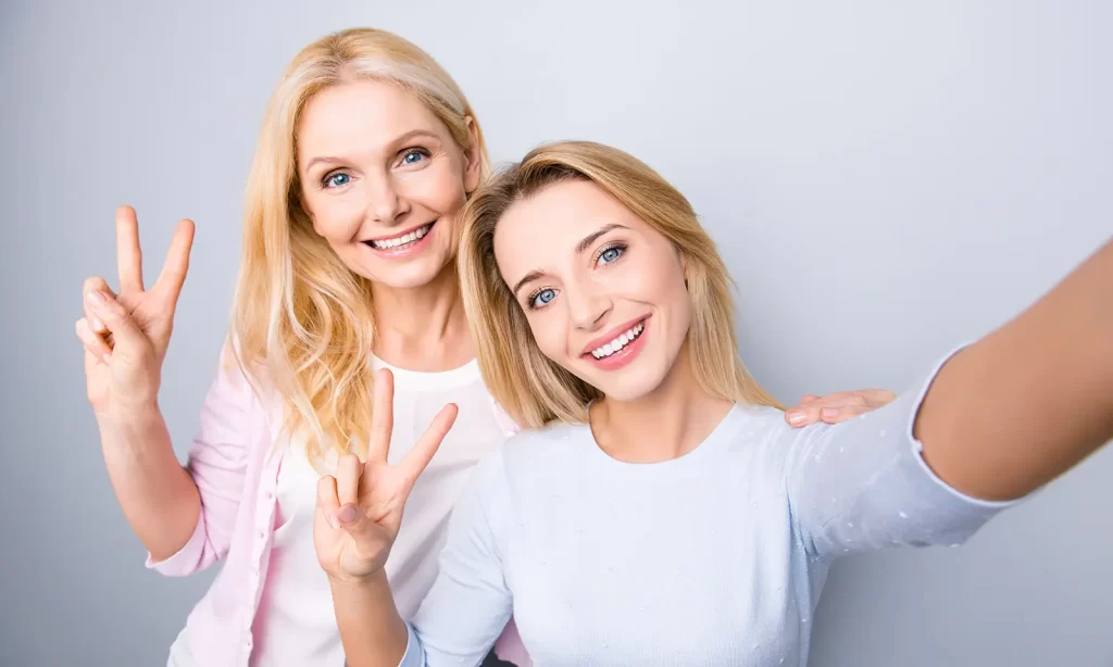 two ladies wearing white and smiling happily together while holding up peace sign taking selfie