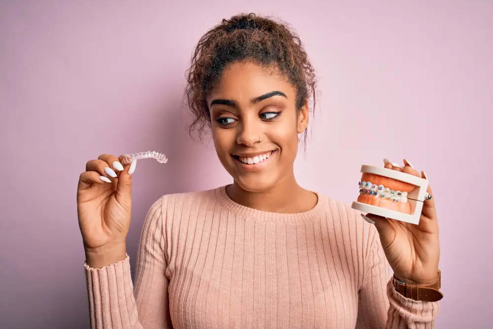 young african american lady smiling and holding invisalign aligners and metal braces typodont