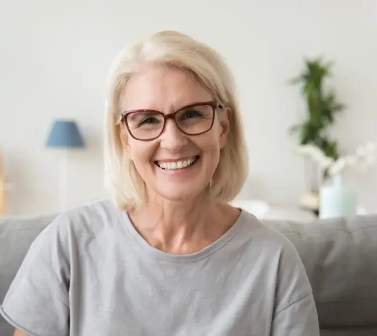 older lady wearing grey shirt and glasses sitting on couch and smiling
