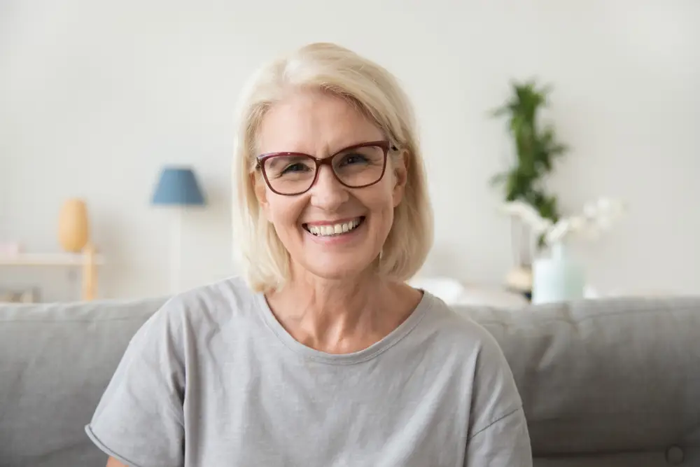 older lady wearing grey shirt and glasses sitting on couch and smiling restorative dentistry duncan