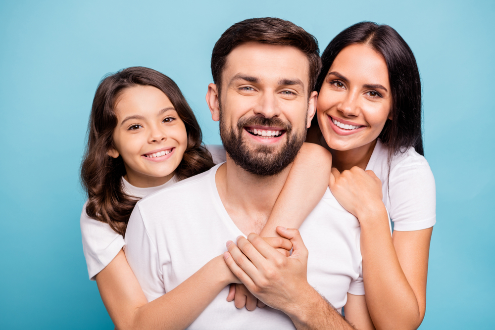 family of three wearing white and smiling together, blue background